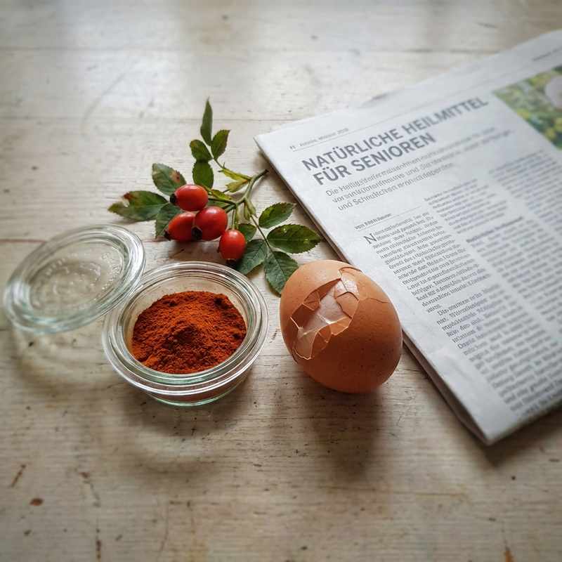 Astaxanthin-Pulver, frische Hagebutten, aufgebrochenes Ei mit Membran auf hellem Holztisch